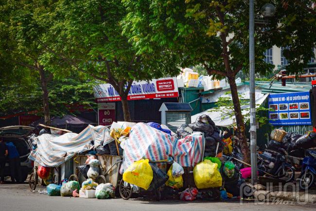 Tại địa điểm thu gom rác thải trên đường Dương Đình Nghệ, tình trạng rác quá tải đang trở thành mối lo lắng của người dân nơi đây. Rác chất đống đầy ắp ngồn ngộn trong thùng xe, công nhân phủ bạt chờ xe đến thu gom. Các hộ gia đình vì không có chỗ đổ rác tiếp tục vứt thêm các túi rác to nhỏ khiến khung cảnh vô cùng mất mỹ quan.